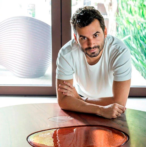 Lucas Recchia veste camiseta branca e está apoiado na mesa "Janelas"