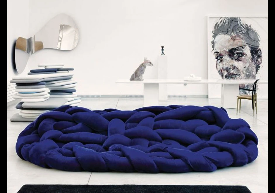 Sofá de design moderno com estrutura tubular preenchida com várias densidades de espuma na cor azul escuro, visto de frente, em uma sala com decorações brancas e cinzas, incluindo um quadro com rosto de mulher e um espelho moderno e curvado.