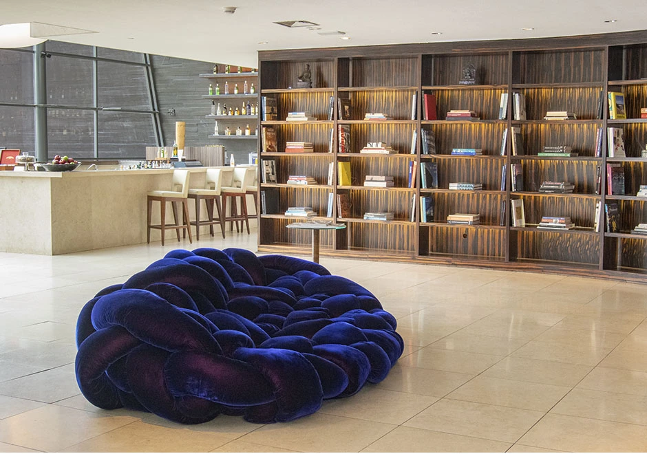 Sofá de design moderno com estrutura tubular preenchida com várias densidades de espuma na cor azul escuro, visto de lado, em uma sala com uma estante de livros e uma bancada com cadeiras altas na cor bege.