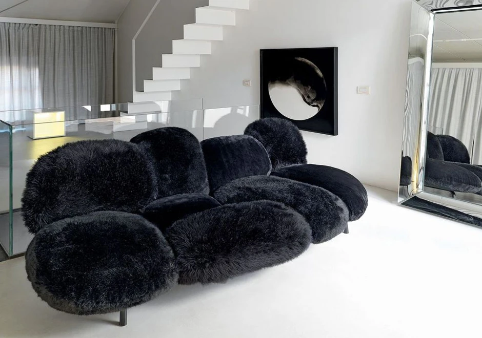 Sofá de design moderno, com estrutura de aço e estofado em pelúcia preto, visto de lado, em uma sala com um espelho, uma escada ao lado direito e umquadro ao fundo.