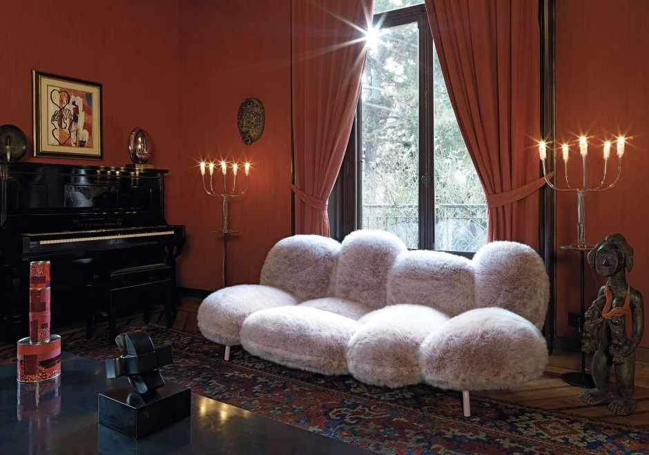 Sofá de design moderno, com estrutura de aço e estofado em pelúcia rosado, visto de lado, em uma sala com candelabros, um piano ao lado direito e uma parede vermelha ao fundo com uma grande janela.