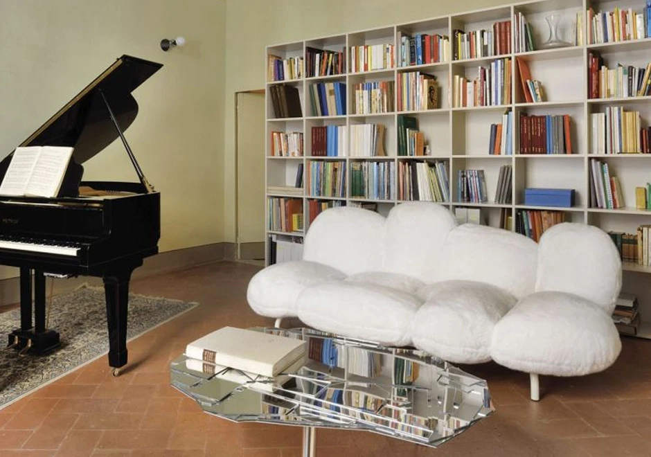 Sofá de design moderno, com estrutura de aço e estofado em pelúcia branco, visto de lado, em uma sala com uma mesa espelhada a frente, um piano ao lado direito e uma estante de livros ao fundo.