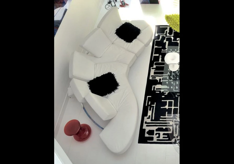 Sofá de design futurista, com estrutura de aço e mecanismo flexível com diferentes inclinações, na cor branca, com almofadas pretas, visto de cima em uma sala moderna.