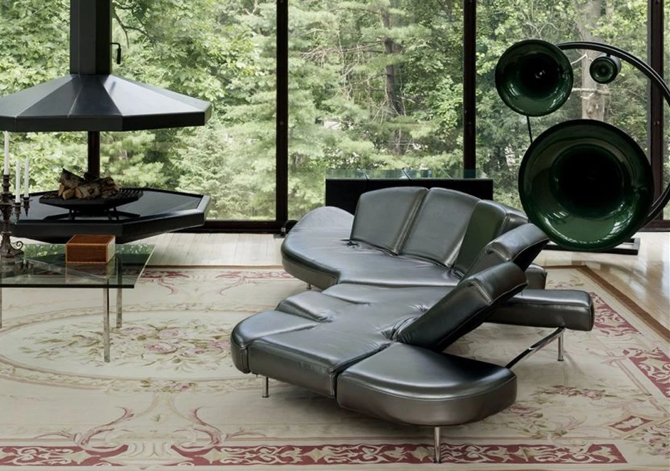 Sofá de design futurista, com estrutura de aço e mecanismo flexível com diferentes inclinações, na cor prata, visto de lado em uma sala moderna com parede de vidro.