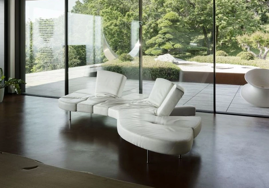Sofá de design futurista, com estrutura de aço e mecanismo flexível com diferentes inclinações, na cor branca, visto de lado em uma sala com parede de vidro e um jardim ao fundo.