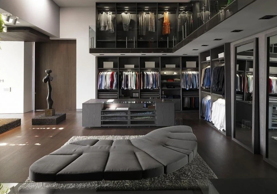 Sofá de design futurista, com estrutura de aço e mecanismo flexível com diferentes inclinações, na cor cinza, visto de lado em um cômodo usado como closet.