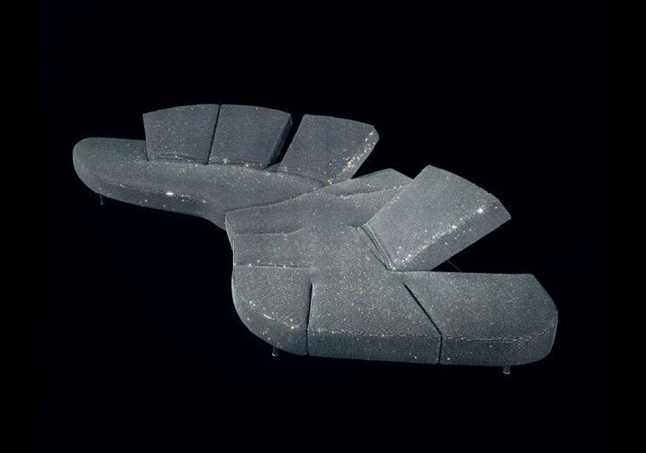 Sofá de design futurista, com estrutura de aço e mecanismo flexível com diferentes inclinações, em tecido tramado com cristais Swarovski, visto de lado em um fundo preto.