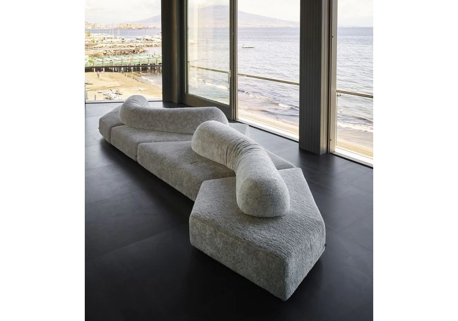 Sofá de design moderno, com pés em ABS, revestido em espuma e com estofado em tecido cinza, visto de lado em uma sala de parede de vidro.