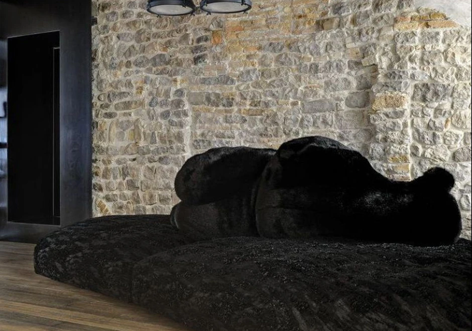 Sofá de design com estrutura de madeira, estofado em plumas e tecido na cor preto, visto de lado, em uma parede de tijolo a vista.
