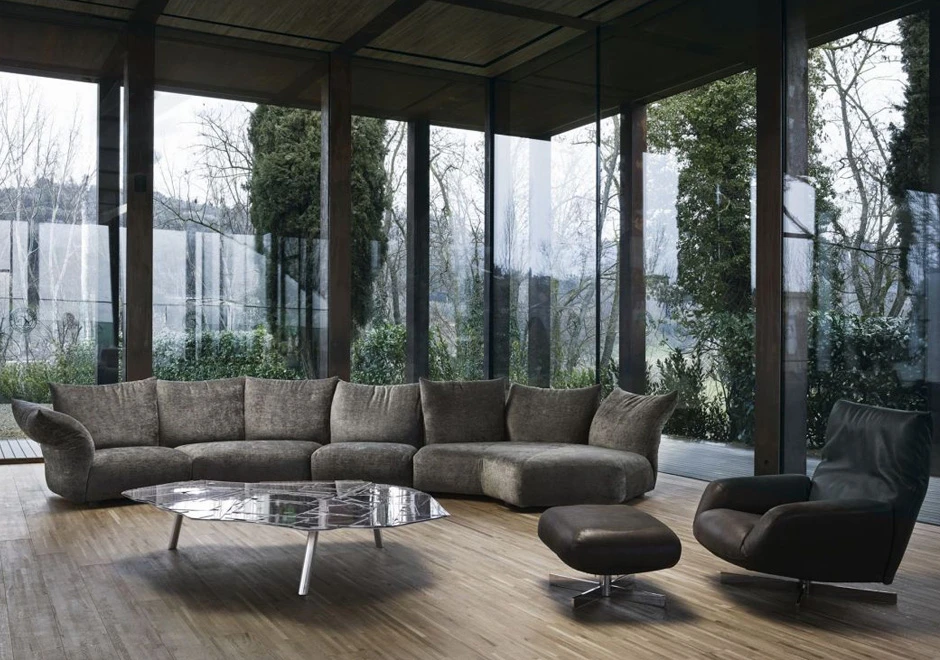 Sofá de design, com estrutura metálica, almofadas e braços reclináveis, cinza, visto de frente em uma sala de parede de vidro.