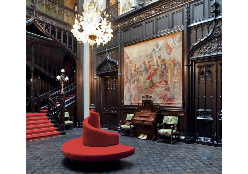 Sofá ilha,com pés em aço cromado e estofado de veludo vermelho, visto de lado em um salão clássico.