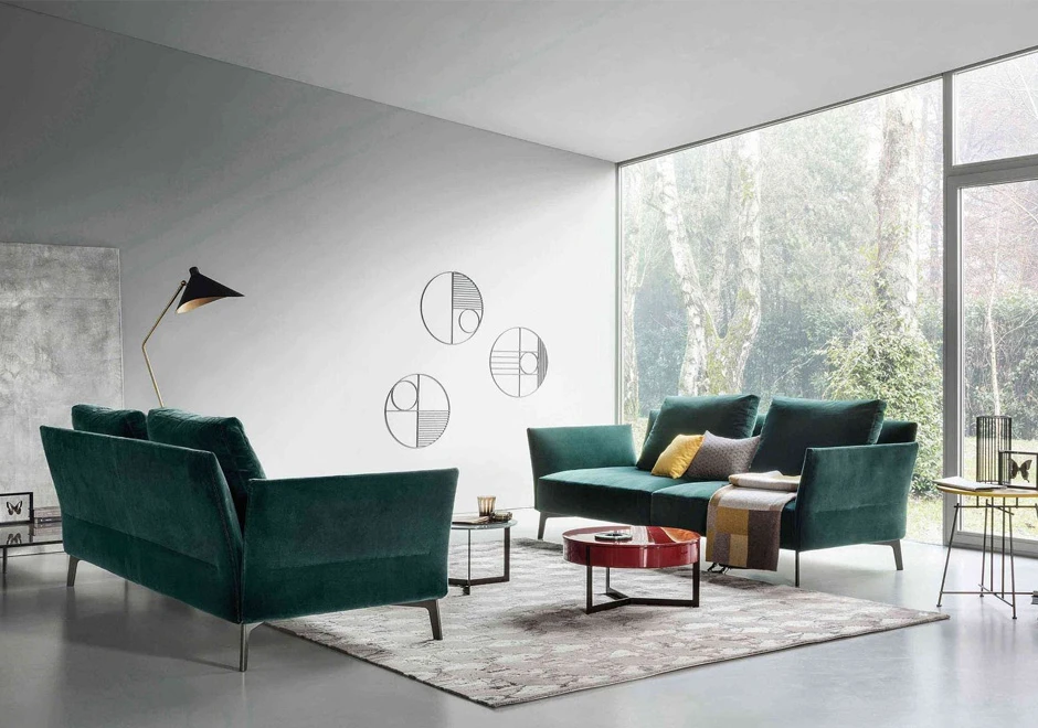 Dois sofás de design clássico, com estrutura de metal e estofado em tecido verde, um de frente para o outro, em uma sala clara, com uma parede de vidro.