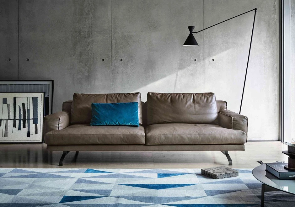 Sofá modular, com estrutura de metal, estofado couro, com uma almofada azul, visto de frente em uma sala moderna.