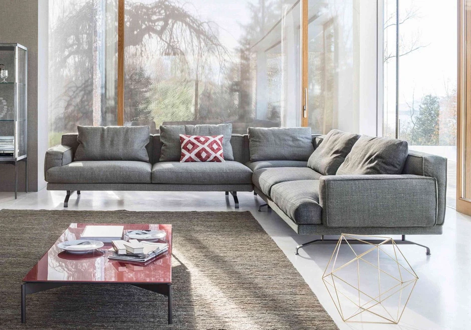 Sofá modular, com estrutura de metal, estofado em tecido cinza e em formato L, visto de frente em uma sala de parede de vidro.