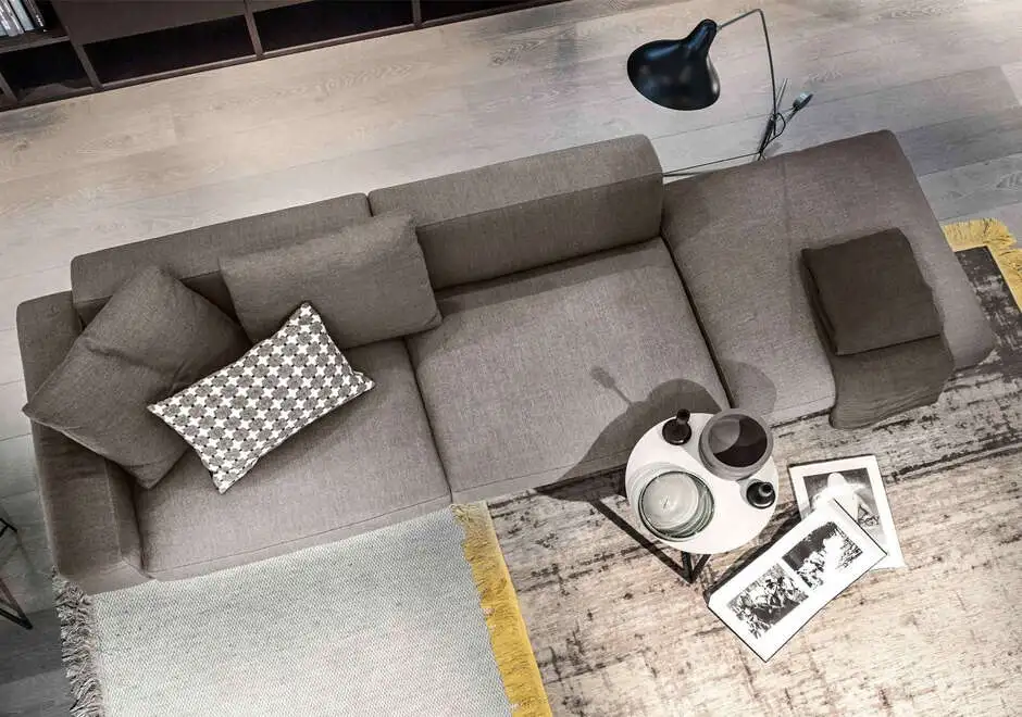 Sofá modular com estrutura de metal, estofado em tecido cinza, visto de cima, em uma sala com estante de livros ao fundo.