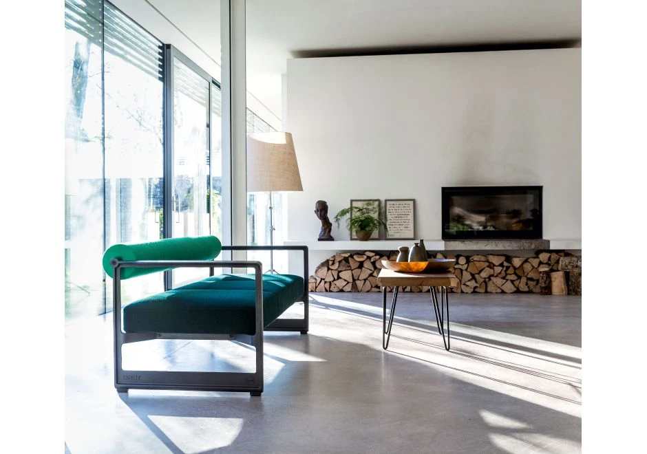 Sofá contemporâneo com estrutura em ferro fundido, e estofado em espuma, verde claro no encosto e verde escuro no assento, visto de lado em uma sala moderna de janelas grandes e sol batendo no interior.