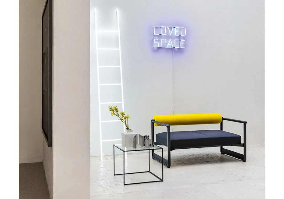 Sofá contemporâneo com estrutura em ferro fundido, e estofado em espuma, amarelo no encosto e azul escuro no assento, visto de lado em uma sala moderna.