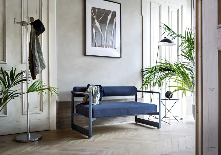 Sofá contemporâneo com estrutura em ferro fundido, e estofado em espuma, azul escuro , visto de lado em uma sala moderna e com uma bolsa em cima.
