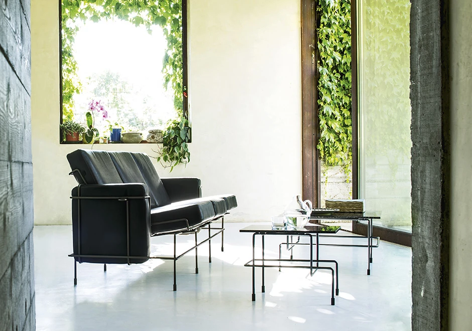 Sofá modular, com estrutura em aço e almofadas em espuma, em tecido cinza, visto de lado em uma sala de parede de vidro.