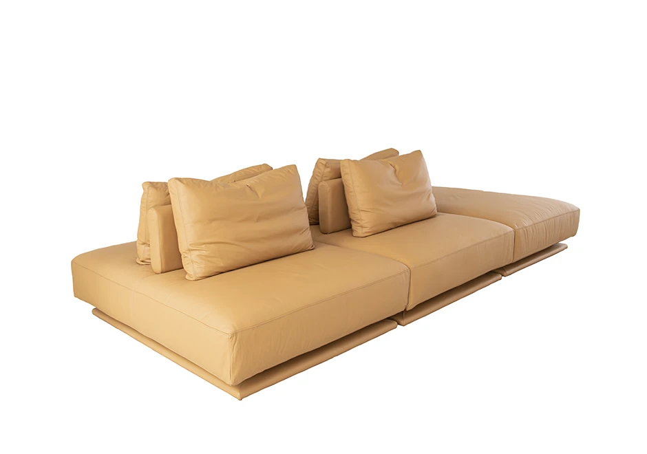 Sofá modular com pés em madeira e revestimento em tecido bege, com quatro almofadas do mesmo material, visto de lado, em um fundo branco.