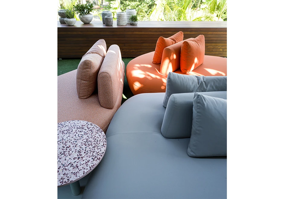 Detalhe de sofá Pedras Outdoor com tecidos azul, rosa e laranja e almofadas decorativas, visto de frente em uma area externa.