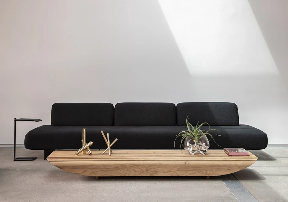 Sófa modular, com base de metal, revestido em espuma, estofado em tecido preto, visto de lado em uma sala com parede branca.