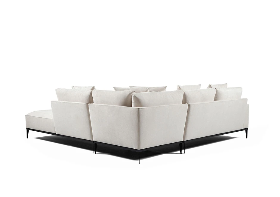 Sofá modular branco, com pés em aço escuro, com diversas almofadas, visto de costas, em formato de L, em um fundo branco