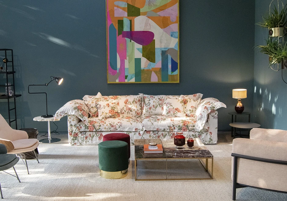 sofá modular com estampa de flores centralizado e visto de frente, com mesinhas ao seu lado e à sua frente, abajures, uma poltrona e duas cadeiras cortadas, um vaso de plantas do lado direito e um quadro colorido ao fundo.