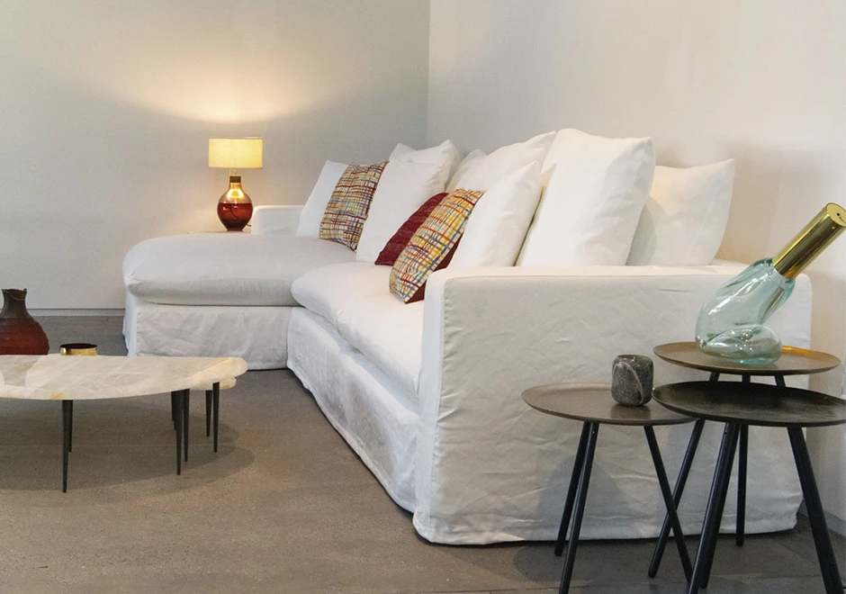 Sofá modular estofado em tecido branco e almofadas decorativas, visto de lado em uma sala de parede branca.