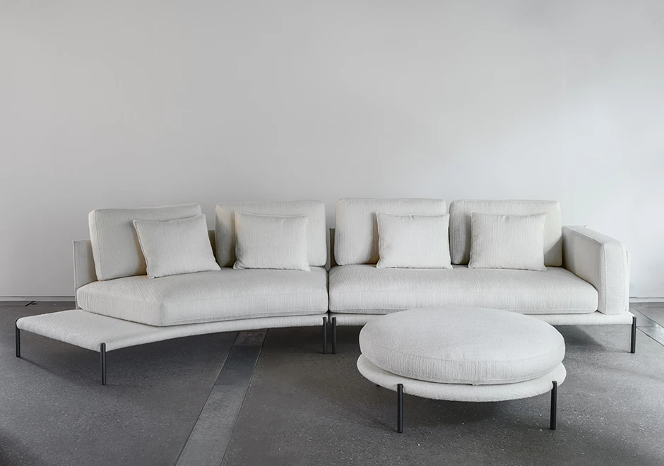 Sofá com escosto com pés de ferro, na cor branca, com um pufe branco, visto de frente com uma parede branca ao fundo.