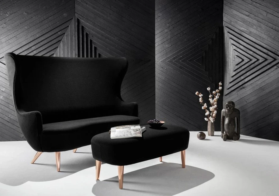 Sofá de design elegante, com estrutura de madeira, estofado em tecido preto, visto de lado com um pufe, em um fundo escuro.