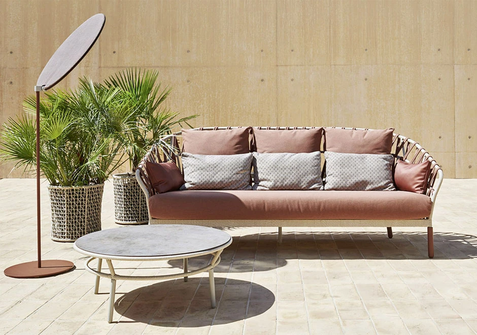 Sofá com encosto em fita trançada à mão, estrutura de alumínio, estofado na cor rosa e almofadas brancas, visto de frente em uma area externa.