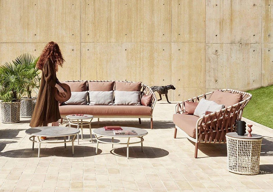 Sofá com encosto em fita trançada à mão, estrutura de alumínio, estofado na cor rosa e almofadas brancas, visto de lado em uma area externa, junto com uma mulher e um cachorro.