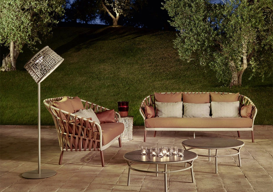 Sofá com encosto em fita trançada à mão, estrutura de alumínio, estofado na cor rosa e almofadas brancas, visto de frente em uma area externa, a noite.