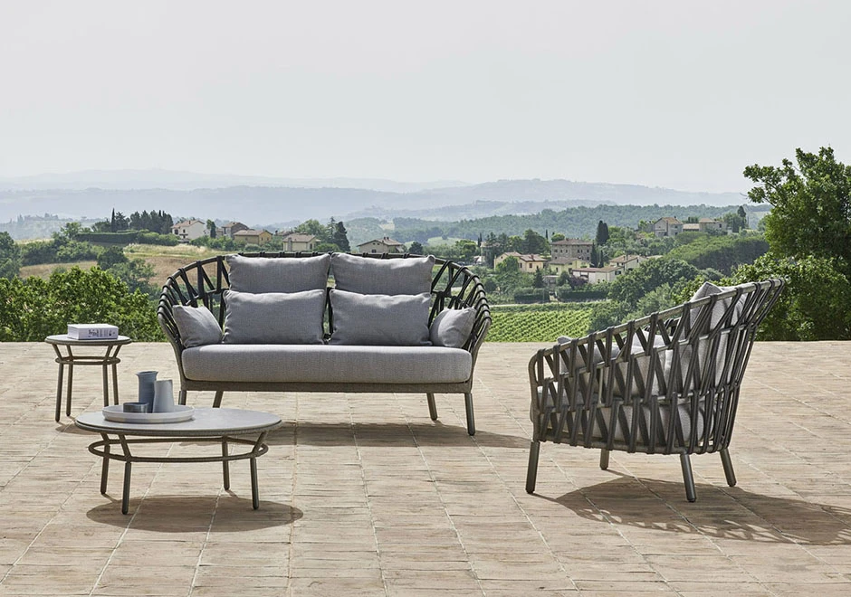 Sofá com encosto em fita trançada à mão, estrutura de alumínio, estofado na cor cinza e almofadas, visto de frente em uma area externa.