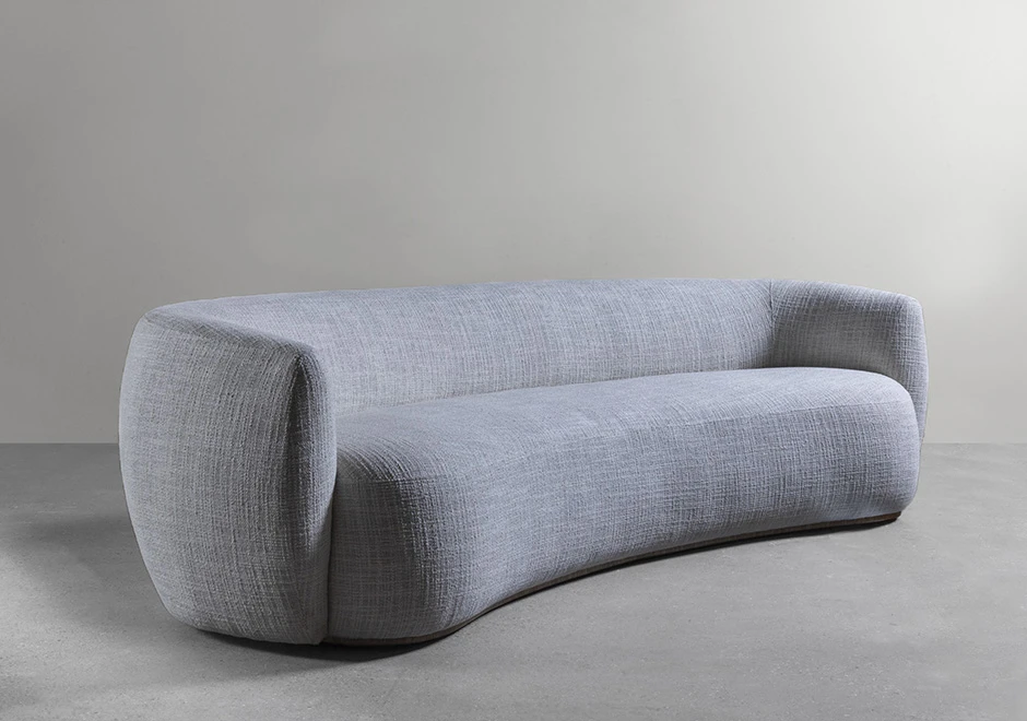 Sofá curvo, estofado em tecido cinza, visto de lado, em um fundo branco.