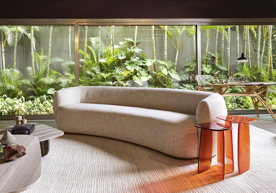 Sofá curvo, estofado em tecido branco, visto de lado em uma sala de parede de vidro com um jardim ao fundo.