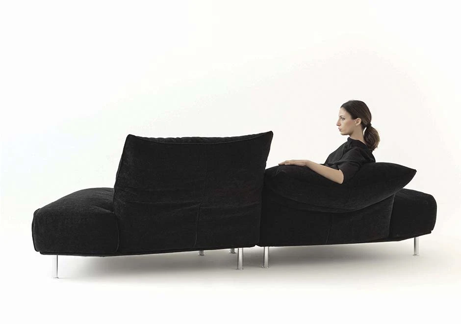 Sofá modular estofado em tecido cinza, visto de costas, com uma pessoa sentada, em um fundo branco.