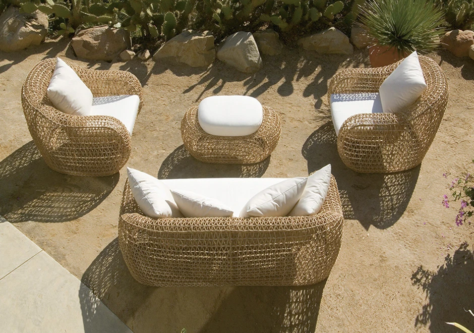 Imagem vista de cima, de um sofá de dois lugares de tecido branco, almofadas brancas e estrutura em fibra sintética trançada, duas poltronas de de tecido branco, almofadas brancas e estrutura em fibra sintética trançada e um pufe ao centro com as mesmas caracteristicas. Imagem em uma área externa de jardim.