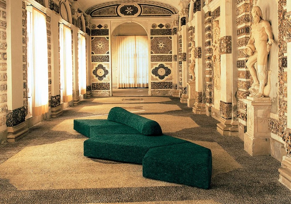 Sofá de design moderno, com pés em ABS, revestido em espuma e com estofado em tecido verde escuro, visto de lado em uma sala com arte clássica.