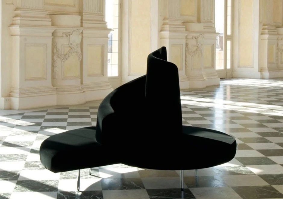 Sofá ilha,com pés em aço cromado e estofado de veludo preto, visto de frente em um salão vazio.