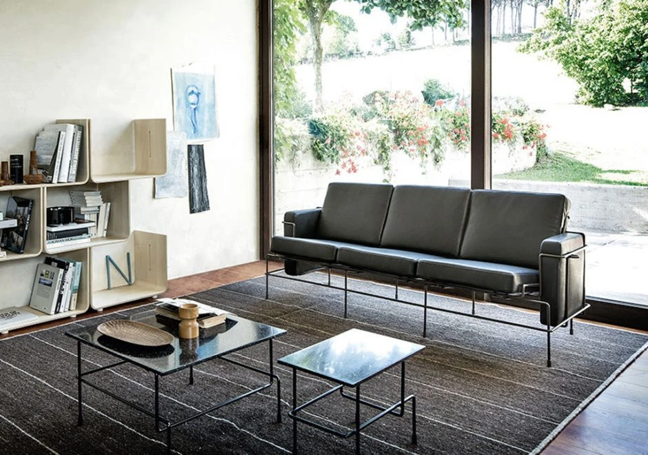Sofá modular, com estrutura em aço e almofadas em espuma, em tecido cinza, visto de frente em uma sala de parede de vidro.