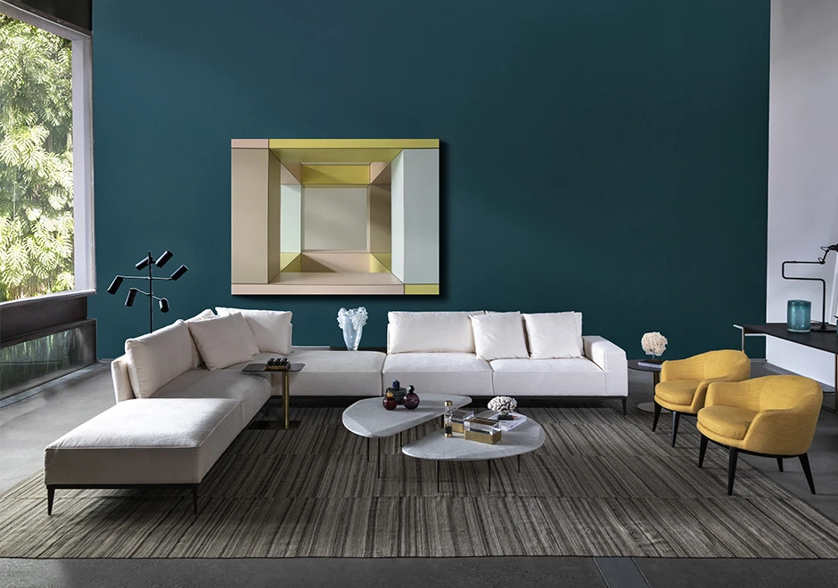 Sofá modular branco, com diversas almofadas, visto de frente, em uma sala de parede azul e janela ampla. Ao seu redor um abajur moderno a esquerda, uma mesa de centro e poltronas amarelas a direita.