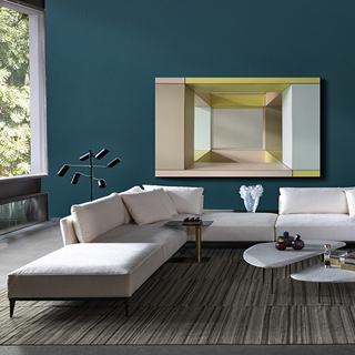 Sofá modular branco, com diversas almofadas, visto de frente, em uma sala de parede azul e janela ampla. Ao seu redor um abajur moderno a esquerda, uma mesa de centro e poltronas amarelas a direita.