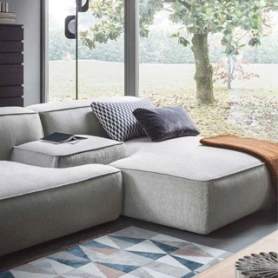 Sofá modular na cor cinza, com duas almofadas , uma cinza e uma xadrez, visto de lado em uma sala com uma parede de vidro ao fundo.