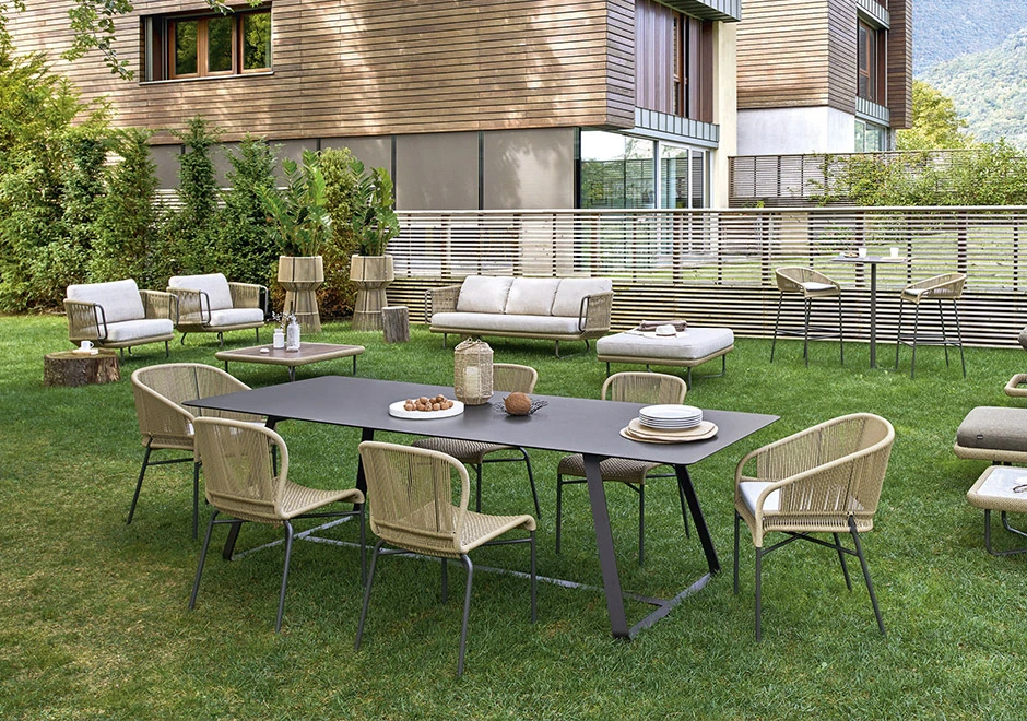 Seis cadeiras assinadas por Anki Gneib com estrutura de alumínio preto e assento bege revestido com fibra sintética trançada a mão, visto de vários angulos, em volta de uma mesa, em um jardim.