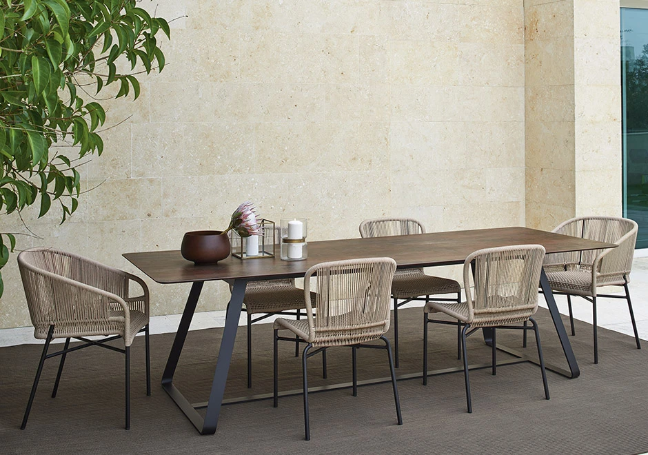 Seis cadeiras assinadas por Anki Gneib com estrutura de alumínio preto e assento bege revestido com fibra sintética trançada a mão, visto de vários angulos, em volta de uma mesa, em uma area externa.