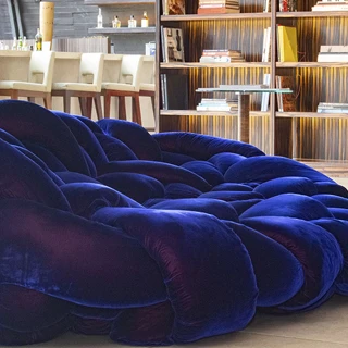 Sofá de design moderno com estrutura tubular preenchida com várias densidades de espuma na cor azul escuro, em uma imagem aproximada, visto de lado, em uma sala com uma estante de livros e uma bancada com cadeiras altas na cor bege.