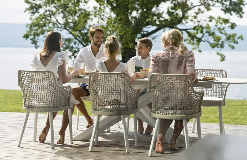 Seis cadeiras assinadas com estrutura em madeira Iroko natural e assento revestido em tecido bege e encosto com estrutura de alumínio com acabamento em corda trançada à mão ou tecido. Visto de frente em uma area externa, com uma família sentada nelas.