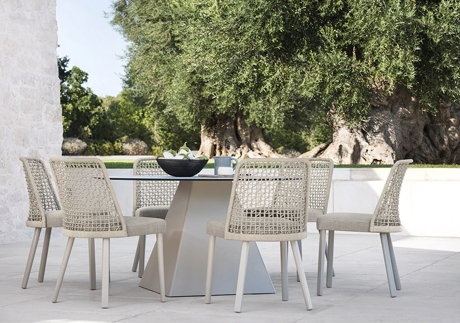 Seis cadeiras assinadas com estrutura em madeira Iroko natural e assento revestido em tecido bege e encosto com estrutura de alumínio com acabamento em corda trançada à mão ou tecido. Visto de frente em uma area externa.